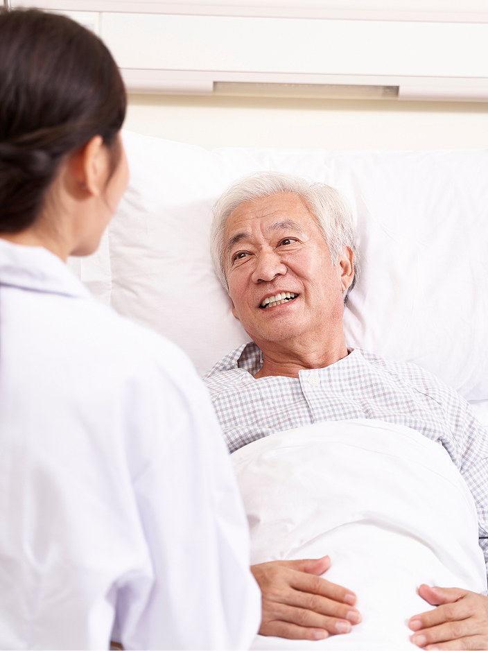 Ein älterer Mann liegt im Krankenhausbett und lächelt eine dunkelhaarige Ärztin an, die vor ihm steht.