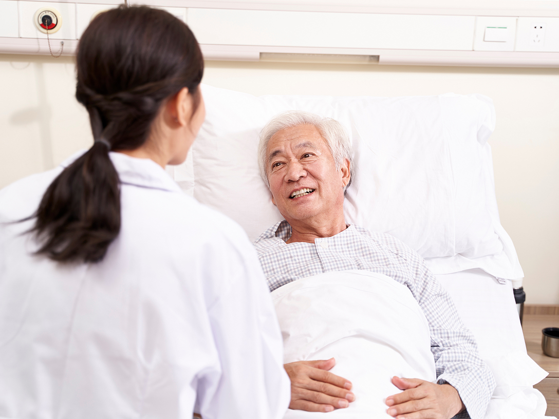 Älterer Mann im Krankenhausbett lächelt eine Ärztin an, die vor seinem Bett steht.