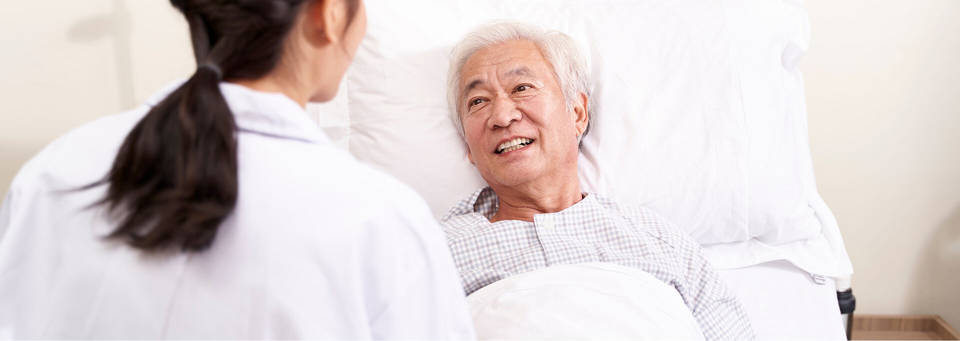 Ein älterer Mann liegt im Krankenhausbett und lächelt eine dunkelhaarige Ärztin an, die vor ihm steht.