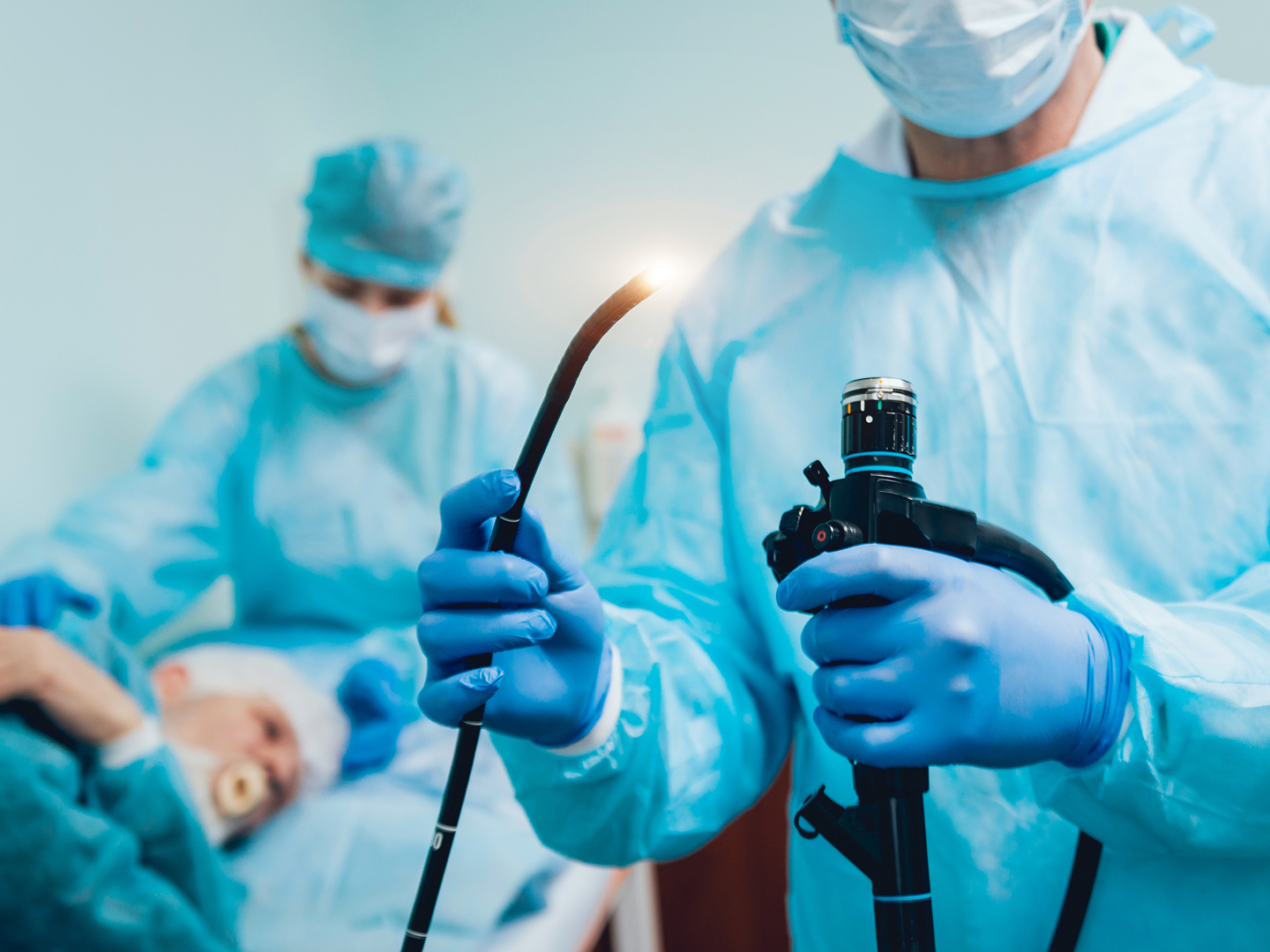 Ein Arzt in Schutzkleidung hält ein medizinisches Gerät mit einer Lichtquelle, während eine Patientin im Hintergrund auf dem OP-Tisch liegt und von einer weiteren Person versorgt wird.