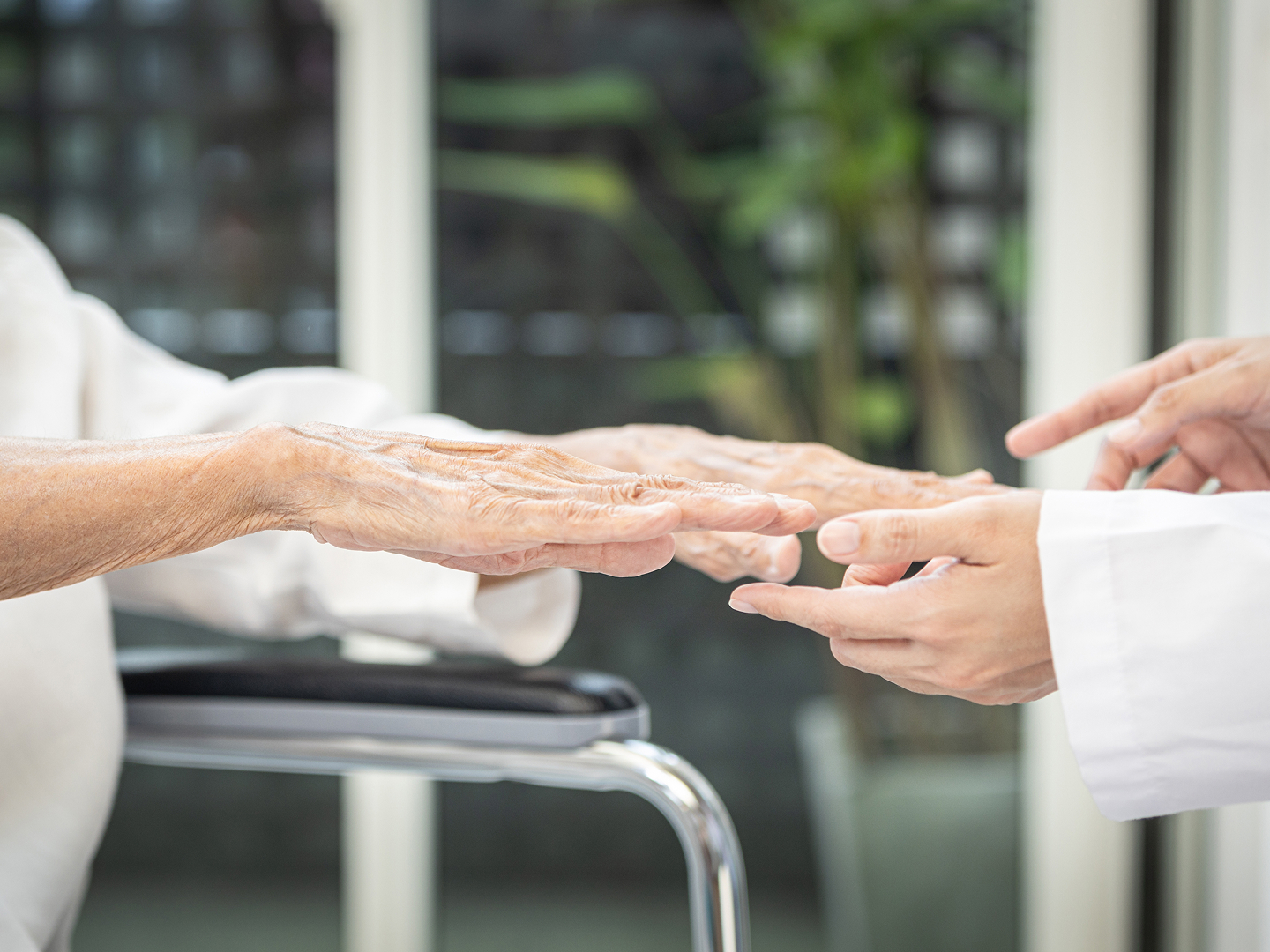 Die Hände einer Patientin im Rollstuhl und die einer medizinischen Fachkraft, die ihr gegenübersteht, berühren sich.