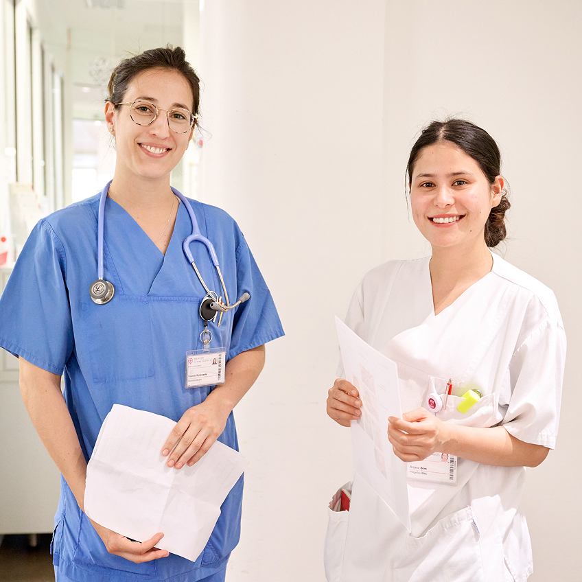 Zwei weibliche, medizinische Fachkräfte in blauer und weißer Uniform stehen lächelnd in einem hellen Flur und halten Dokumente in der Hand.