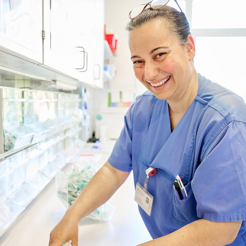 Lächelnde Person in blauer Arbeitskleidung, die an einem Tisch mit medizinischen Materialien arbeitet.