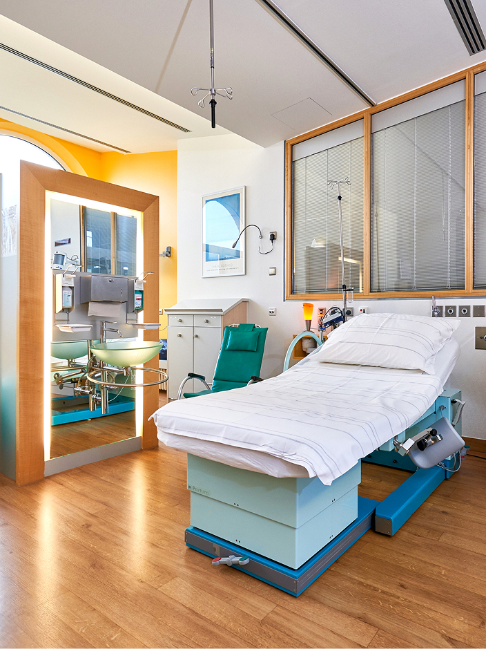 Ein Krankenhauszimmer mit einem Bett, einem Stuhl und einem großen Fenster, das viel Licht hereinlässt.