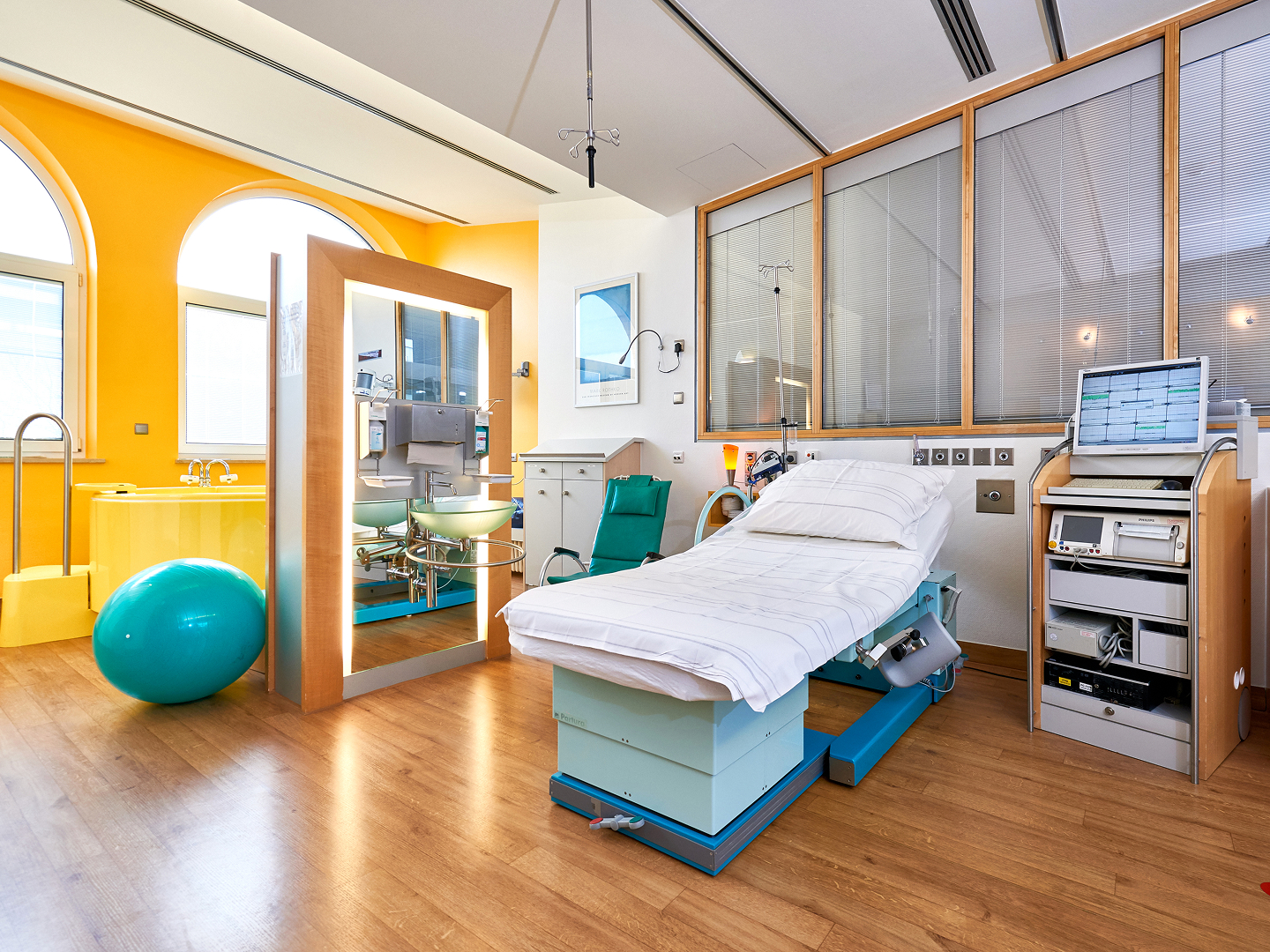 Helles Gebärzimmer mit einem Bett, einem Spiegel, einem grünen Gymnastikstuhl und medizinischen Geräten.