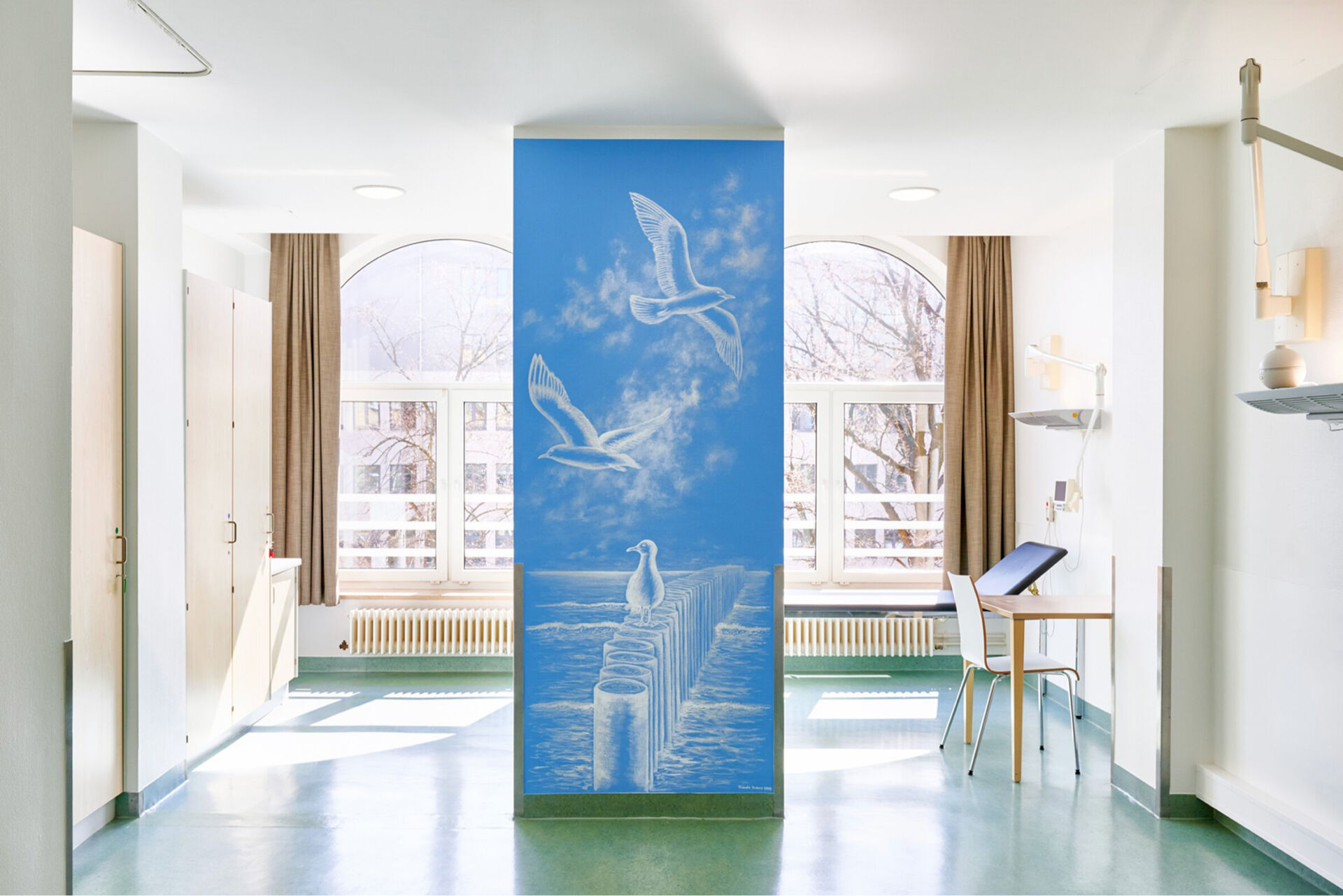 Heller Innenraum mit Stühlen und Fenstern im Hintergrund und einem blauen Wandbild, auf dem Möwen abgebildet sind.