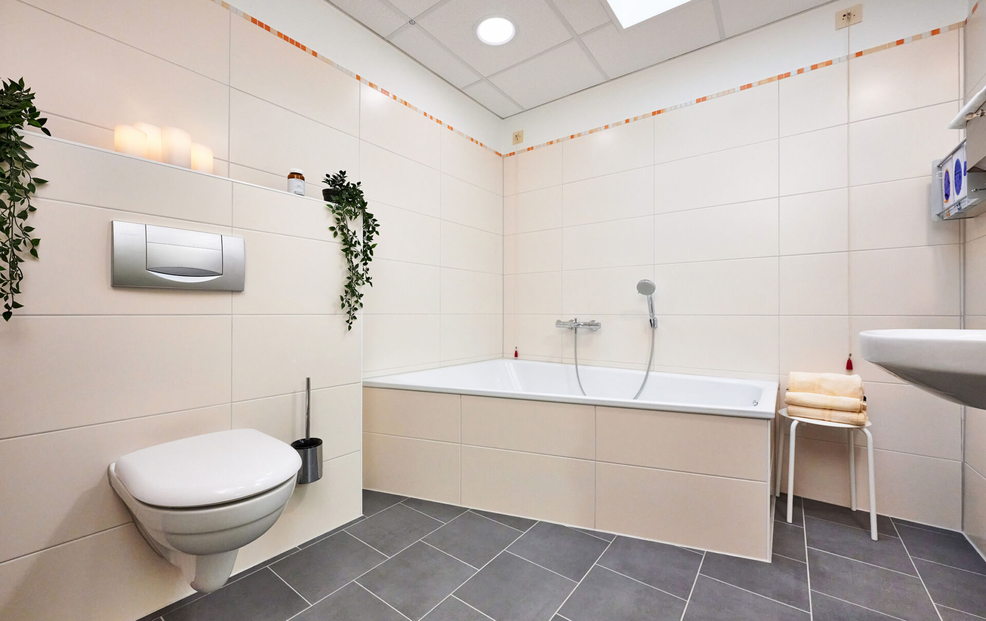 Helles Badezimmer mit Badewanne, Toilette, Waschbecken und grauem Fliesenboden. Pflanzen dekorieren die Wände.