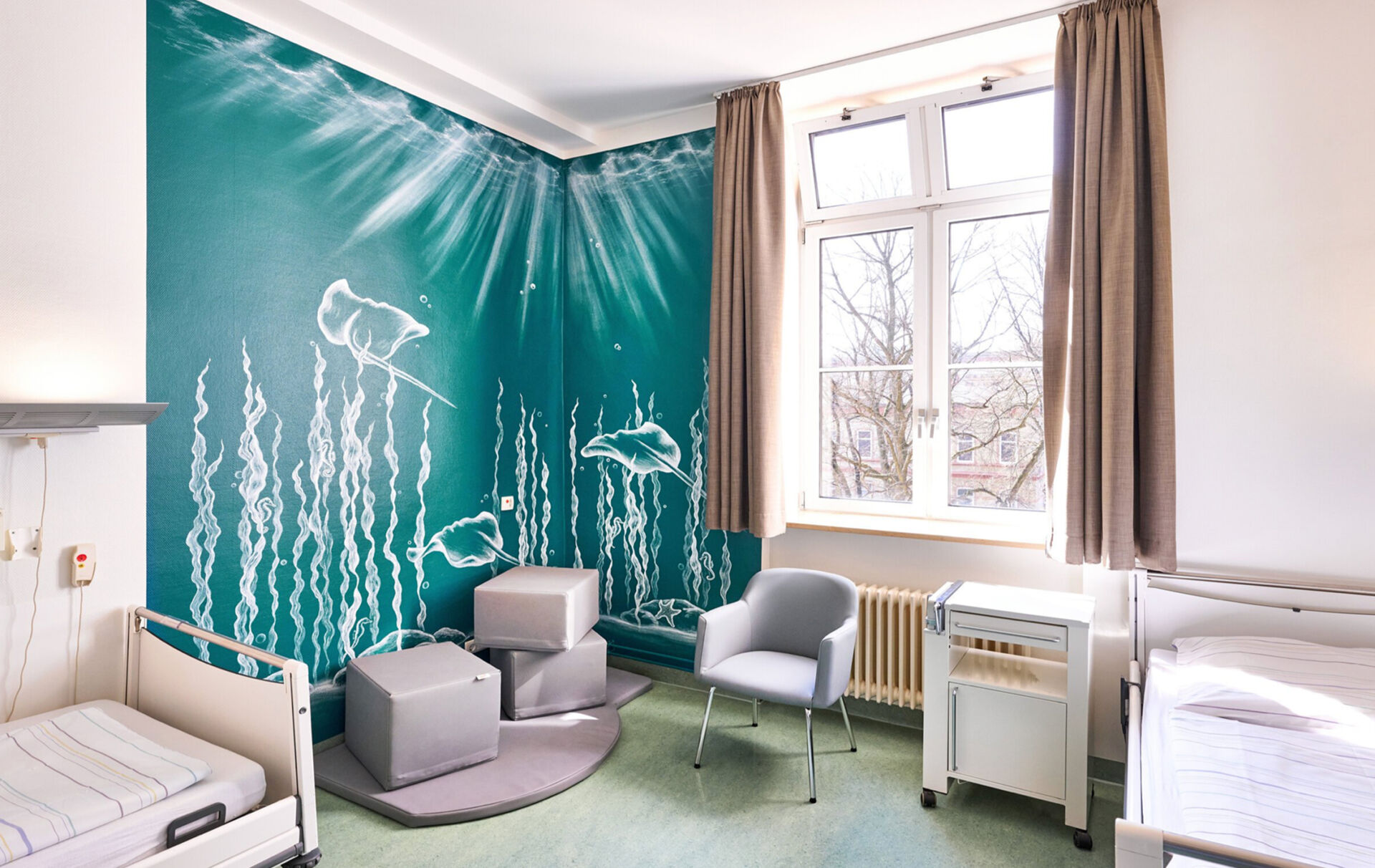 Helles, modernes Patientenzimmer mit zwei Betten, grauen Möbeln und einer Wand mit maritimem Motiv.