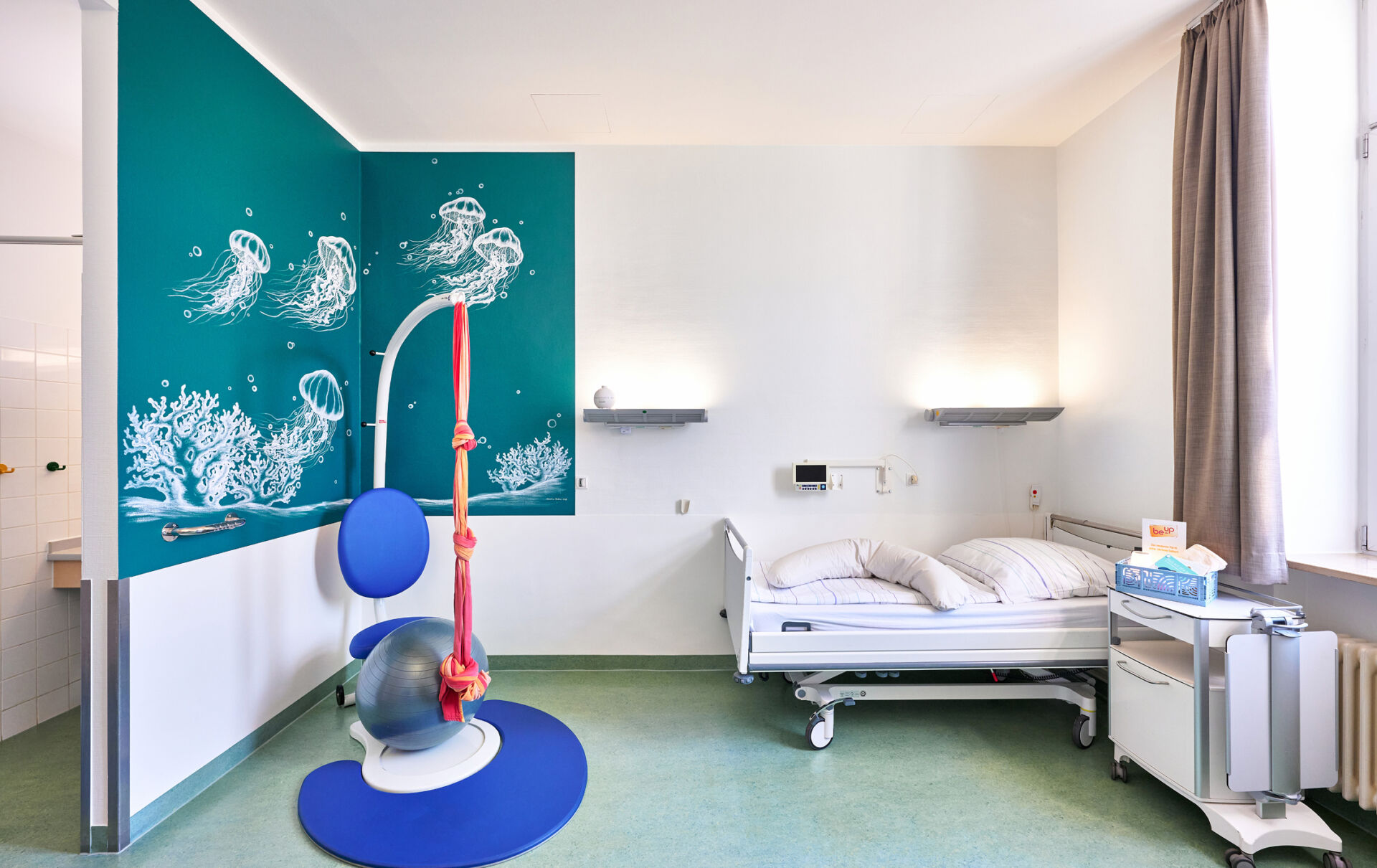 Krankenhauszimmer mit einem Bett, einer bunten Wandgestaltung und einem speziellen Gebärstuhl für Patientinnen.
