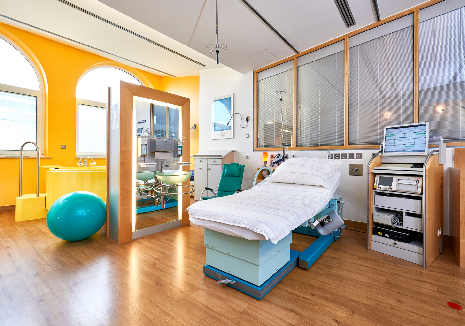 Helles Gebärzimmer mit einem Bett, einem Spiegel, einem grünen Gymnastikstuhl und medizinischen Geräten.