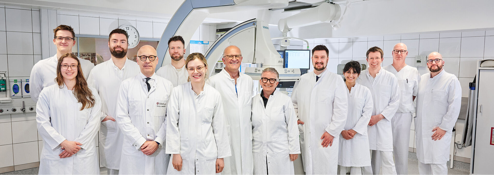 Gruppe von Personen in weißen Kitteln steht in einem modernen Raum mit radiologischen Geräten im Hintergrund.