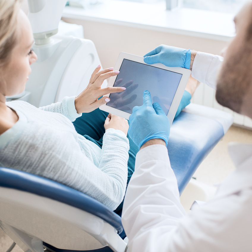Arzt zeigt Patientin auf einem Tablet Informationen, während sie auf einem Behandlungsstuhl sitzt.