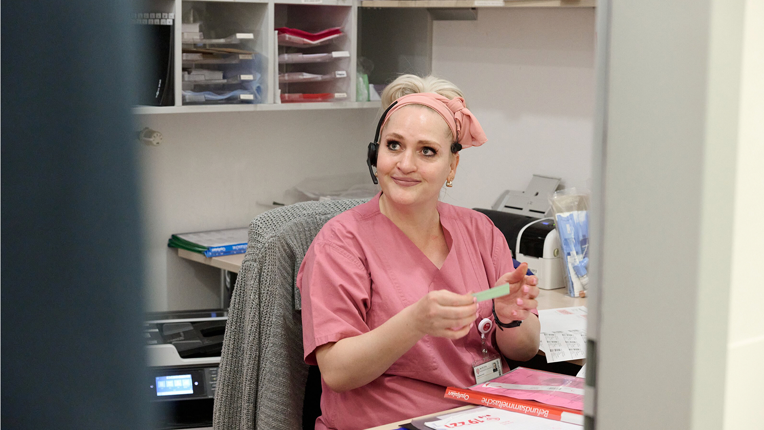 Eine Krankenschwester in rosafarbener Arbeitskleidung sitzt mit Headset an einem Schreibtisch und hält eine Karte in der Hand.