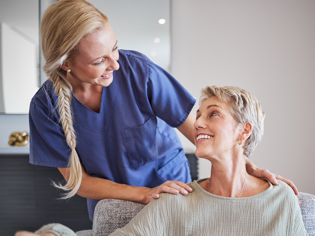 Weibliche Pflegekraft beugt sich von hinten zu einer lächelnden Patientin hinunter und spricht sie freundlich an.