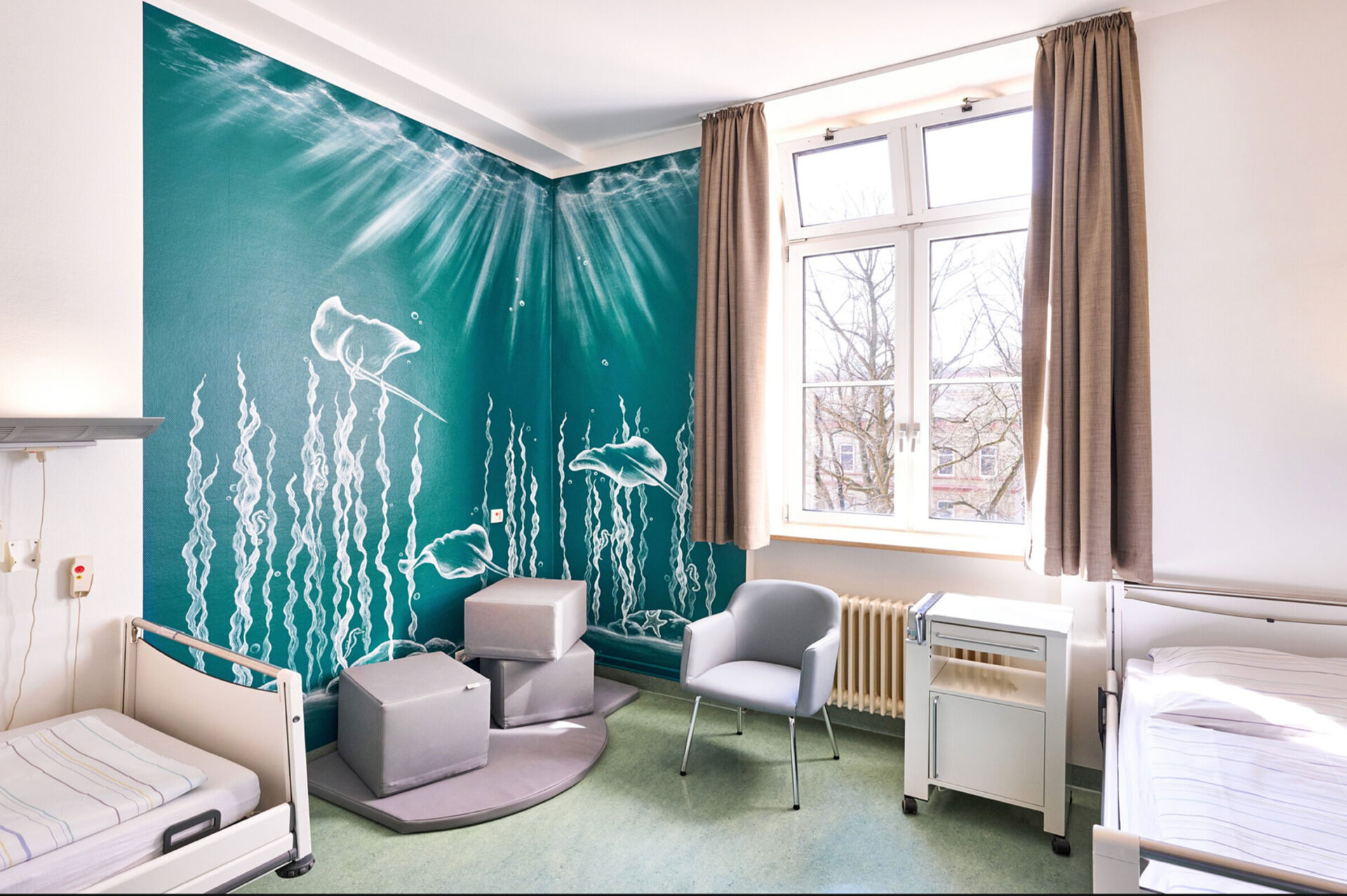 Patientenzimmer mit Fenster, zwei Betten, grauen Sitzmöbeln und einer Wand mit Unterwasser-Motiv.