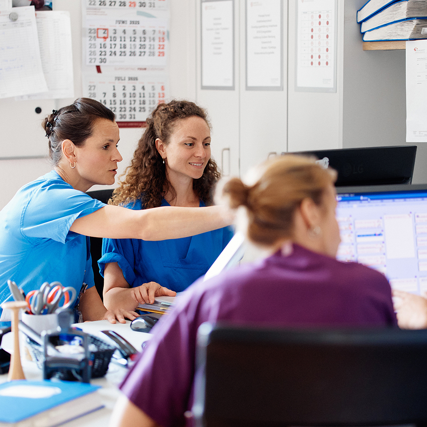 Drei Personen in Arbeitskleidung arbeiten an einem Computer, während eine Person auf den Bildschirm zeigt.