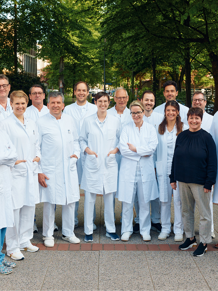 Gruppe von 15 Personen in weißen Kitteln, die im Freien stehen, umgeben von Bäumen und Grünflächen.