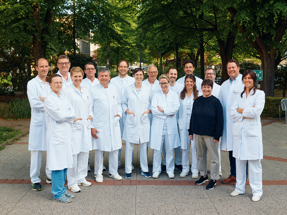 Gruppe von medizinischem Personal in Arbeitskleidung, steht in einem Park, umgeben von Bäumen und Grünflächen.