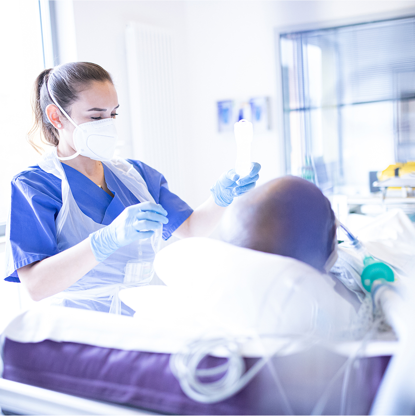Eine medizinische Fachkraft mit Schutzkleidung und Maske behandelt einen Patienten in einem hellen Raum.