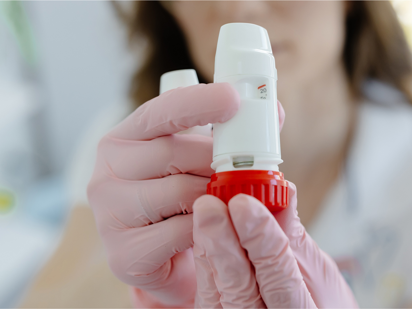 Eine Person mit rosa Handschuhen hält einen weißen Inhalator mit rotem Deckel.