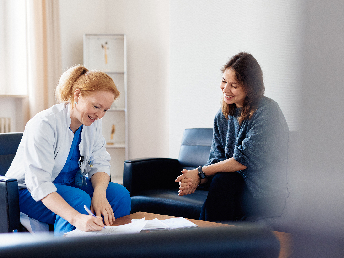 Eine Ärztin sitzt an einem Tisch und schreibt, während eine Patientin ihr gegenüber sitzt und zuschaut.