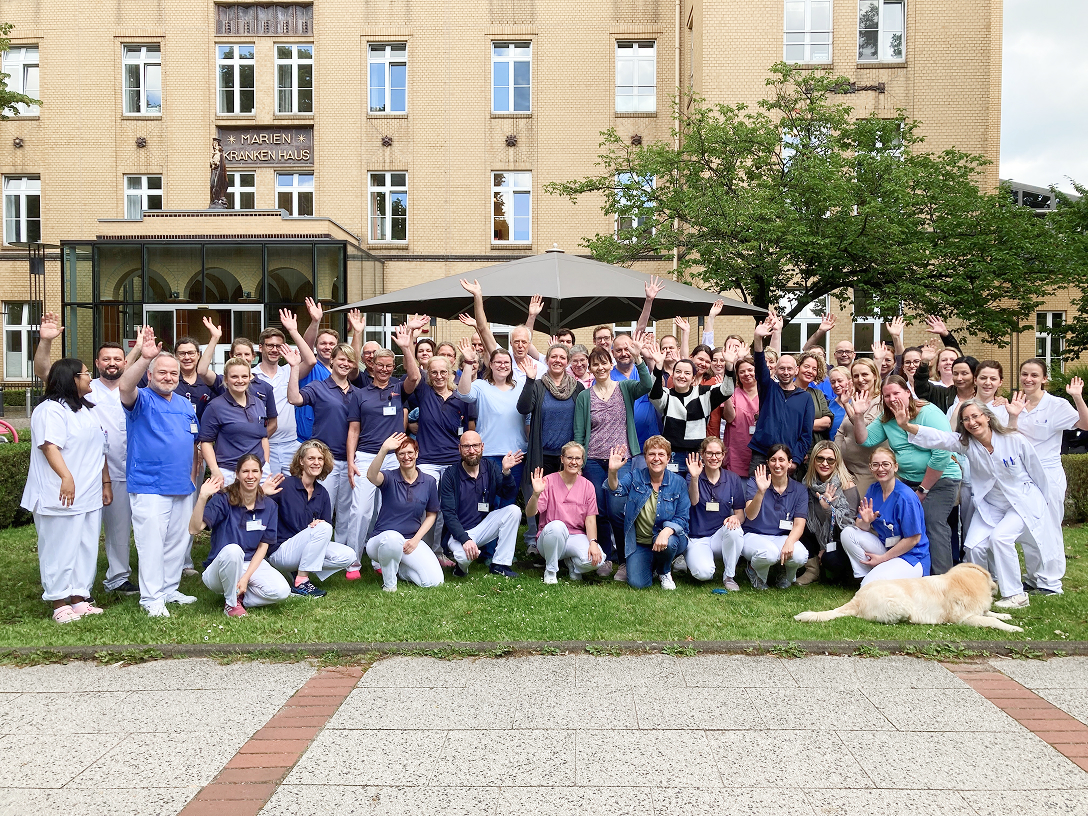 Gruppe von Menschen in Arbeitskleidung, die im Freien stehen und fröhlich in die Kamera winken.