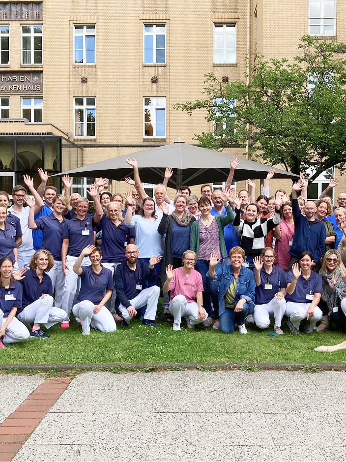 Gruppe von Menschen in Arbeitskleidung, die im Freien stehen und fröhlich in die Kamera winken.