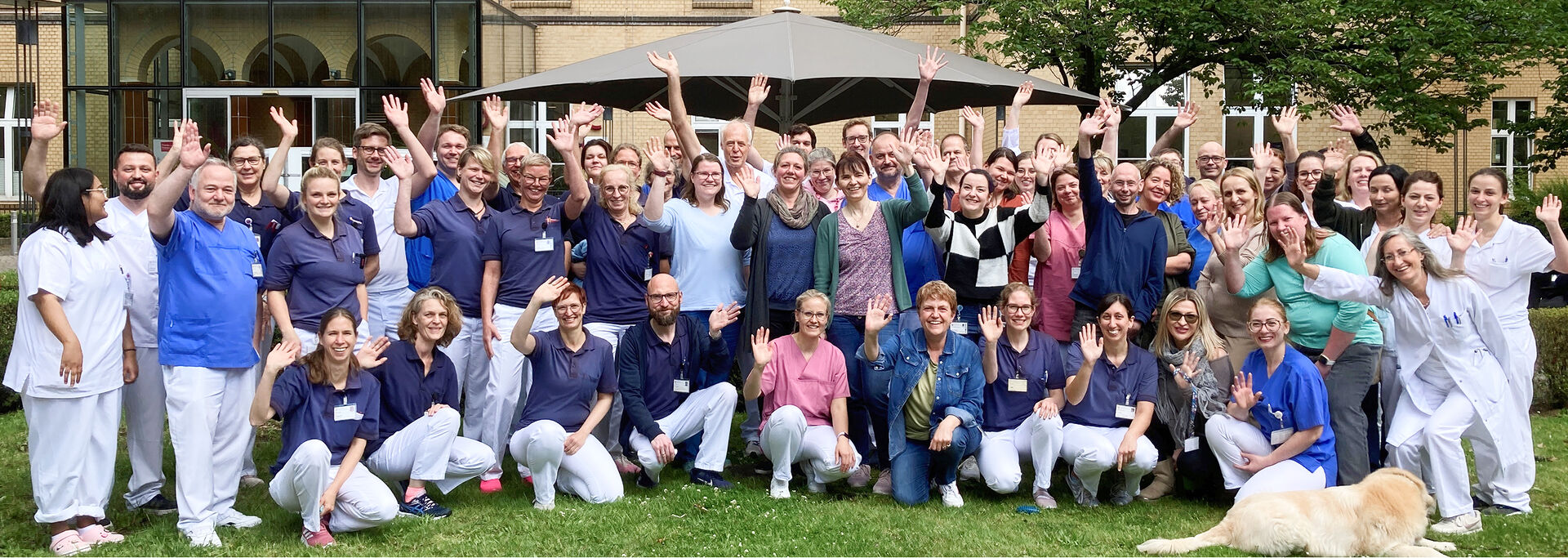 Gruppenshot von über 50 Personen im Freien, die fröhlich die Hände heben, im Hintergrund sind Bäume und ein Gebäude zu sehen.