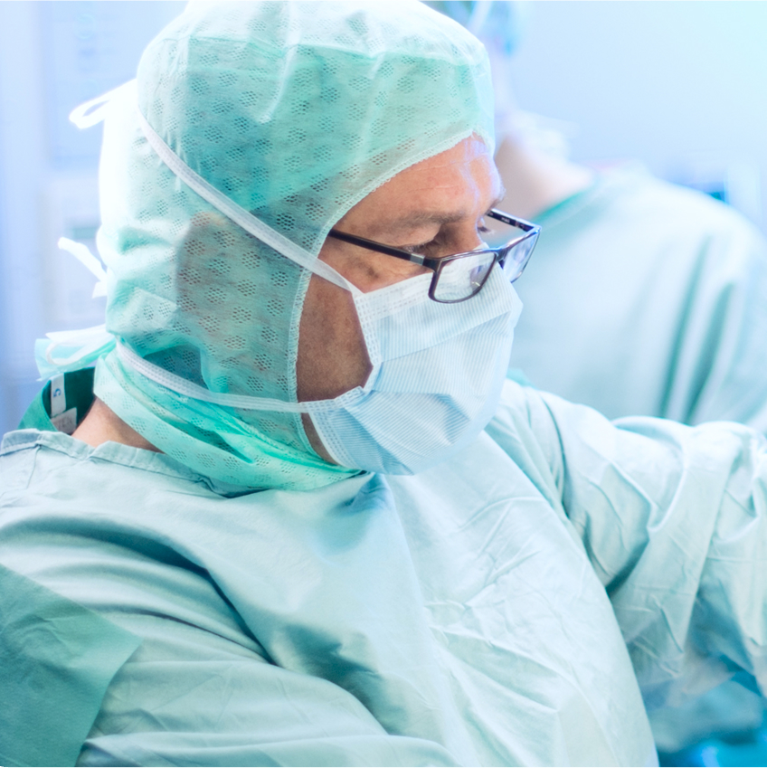 Ein Arzt in OP-Kleidung, mit Maske und Brille, konzentriert bei einer medizinischen Tätigkeit in einem Operationssaal.