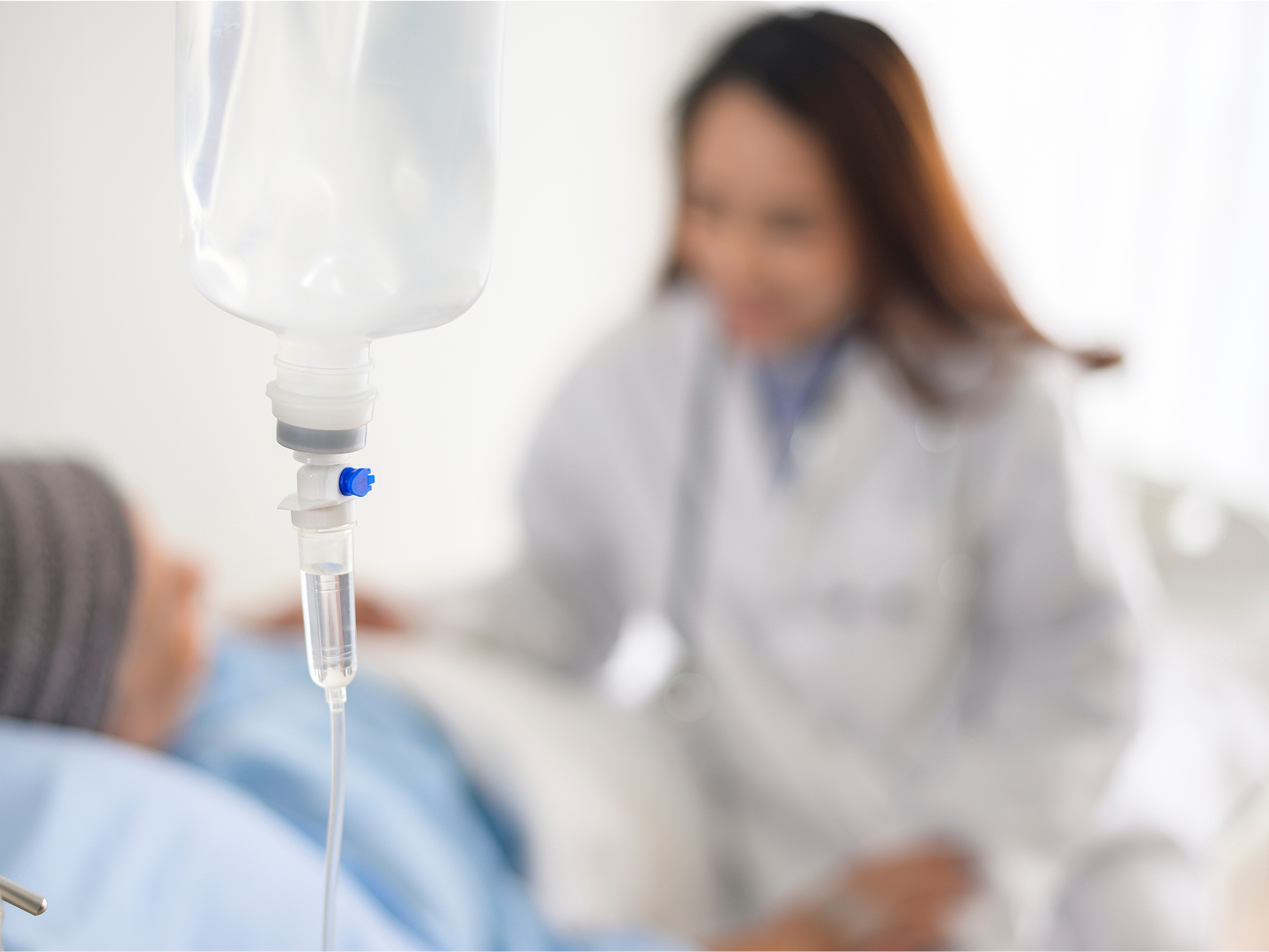 Infusionsbeutel mit Tropfleitung im Vordergrund, im Hintergrund eine Ärztin, die sich um einen Patienten kümmert.
