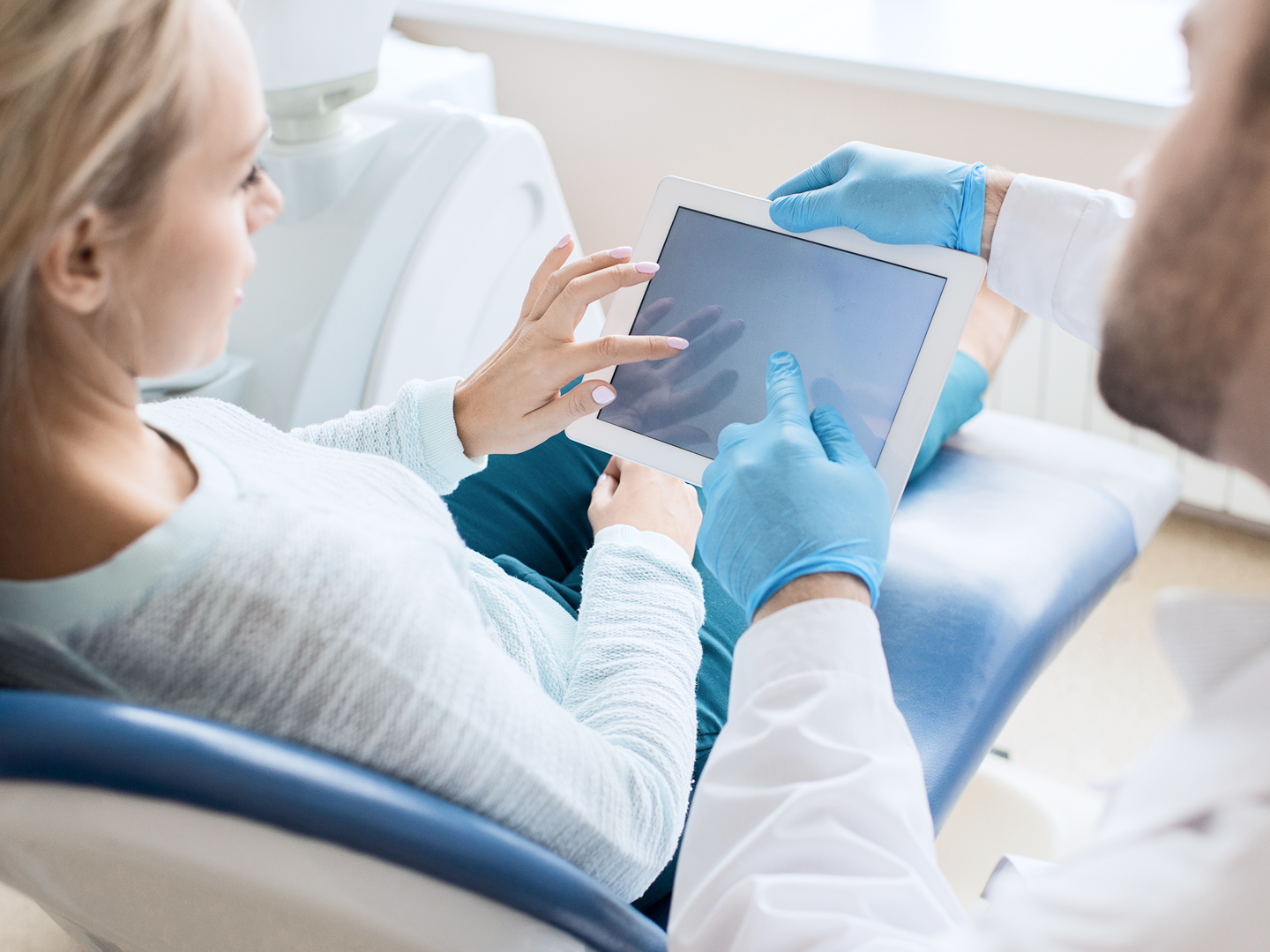 Arzt zeigt Patientin auf einem Tablet Informationen, während sie auf einem Behandlungsstuhl sitzt.