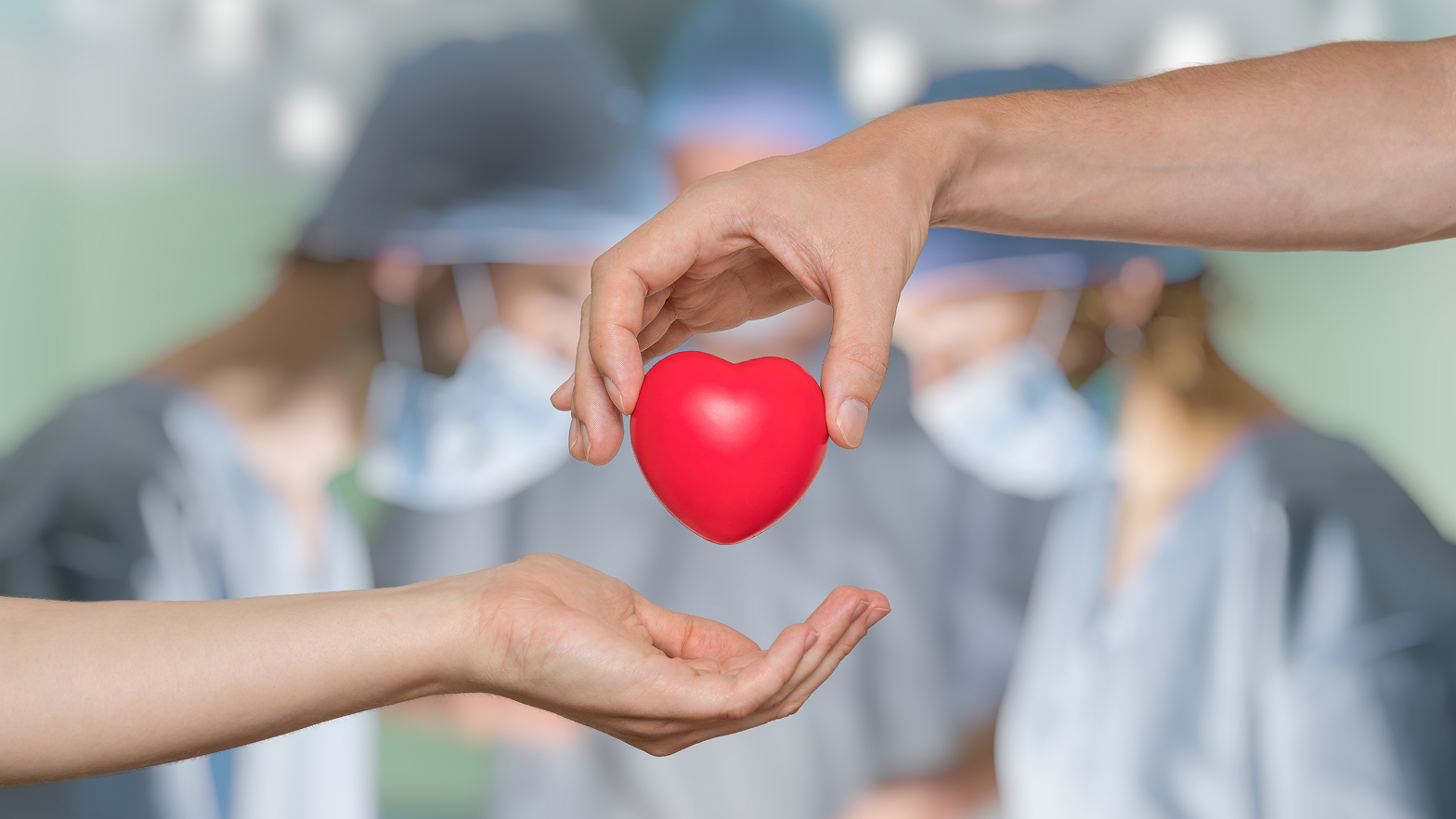 Eine Hand übergibt ein rotes Herz einer anderen Hand, im Hintergrund sind Personen in medizinischer Kleidung zu sehen.