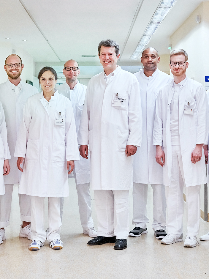 Eine Gruppe von sechs Personen in weißen Kitteln steht in einem Krankenhausflur und lächelt in die Kamera.
