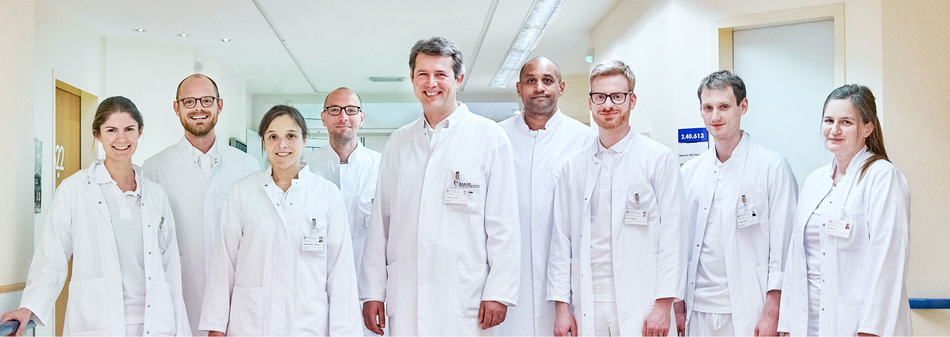 Gruppe von Ärzten in weißen Kitteln, die in einem Krankenhausflur stehen und in die Kamera lächeln.