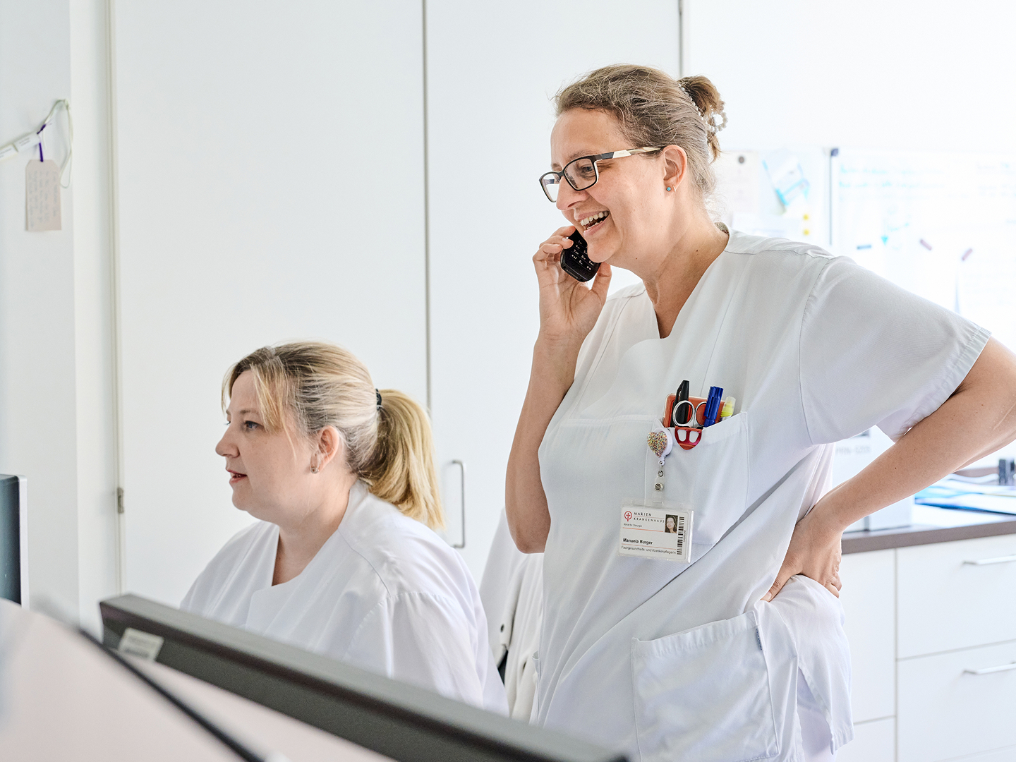 Lächelnde Krankenschwester steht telefonierend hinter dem Empfangstresen, während eine Kollegin neben ihr am Computer arbeitet. Beide tragen weiße Arbeitskleidung.