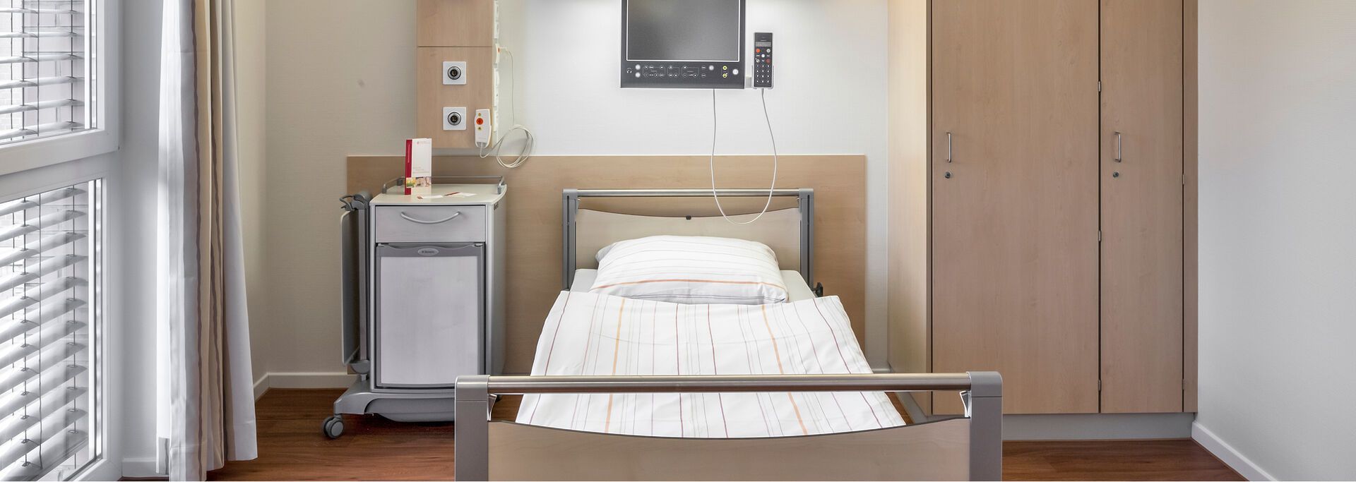 Ein modernes, helles Krankenhauszimmer mit einem Bett, einem Schrank, einem Nachttisch und einem Monitor an der Wand.