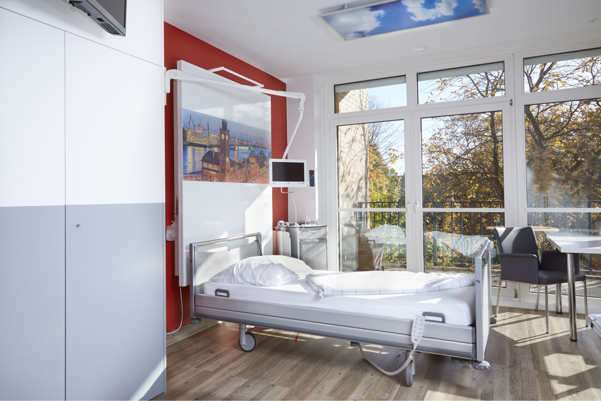 Helles Krankenhauszimmer mit einem Bett, großen Fenstern und einem Tisch mit Stühlen.