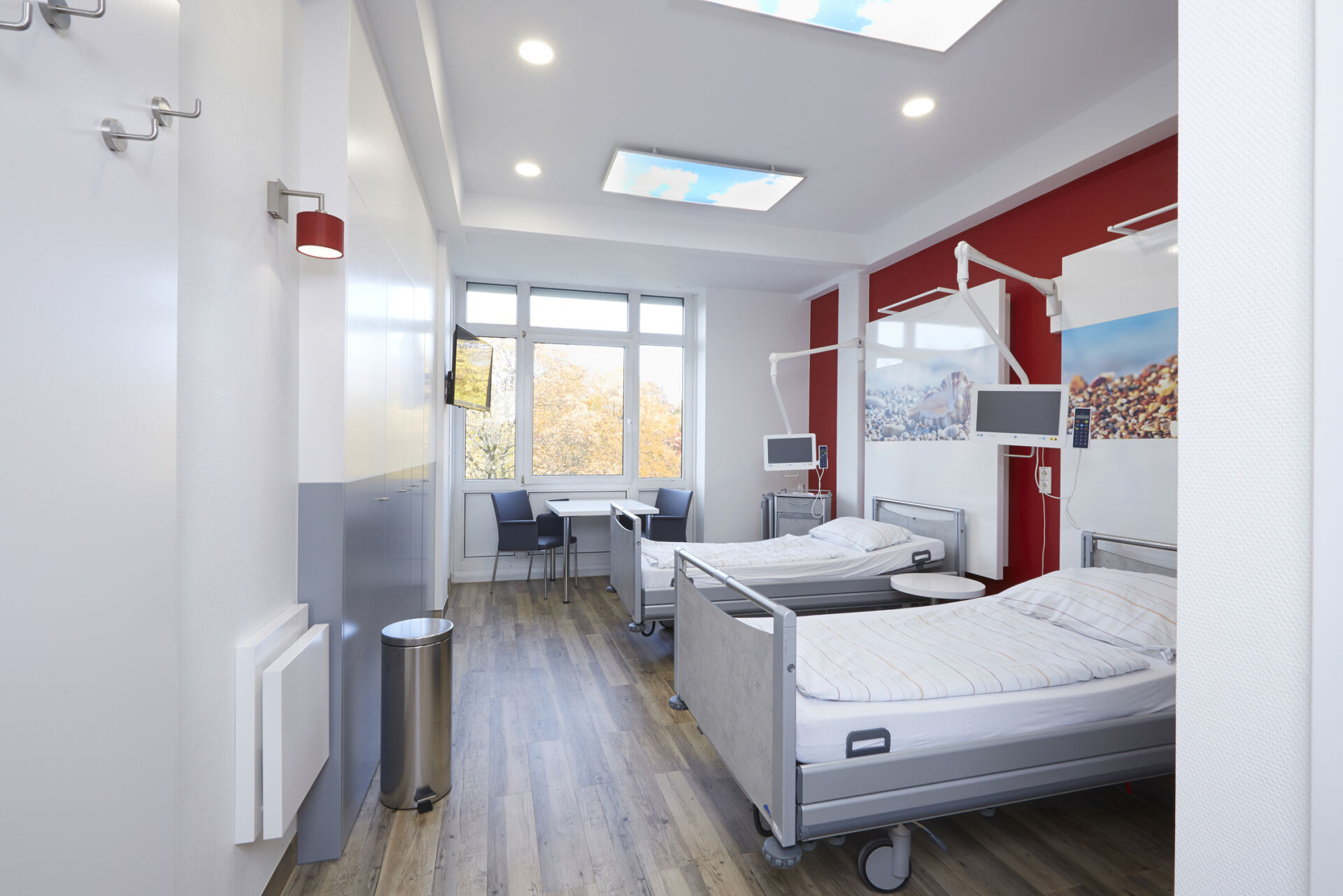 Modernes Krankenhauszimmer mit zwei Betten, Tisch, Stühlen und großem Fenster. Helle Beleuchtung und freundliche Farben.