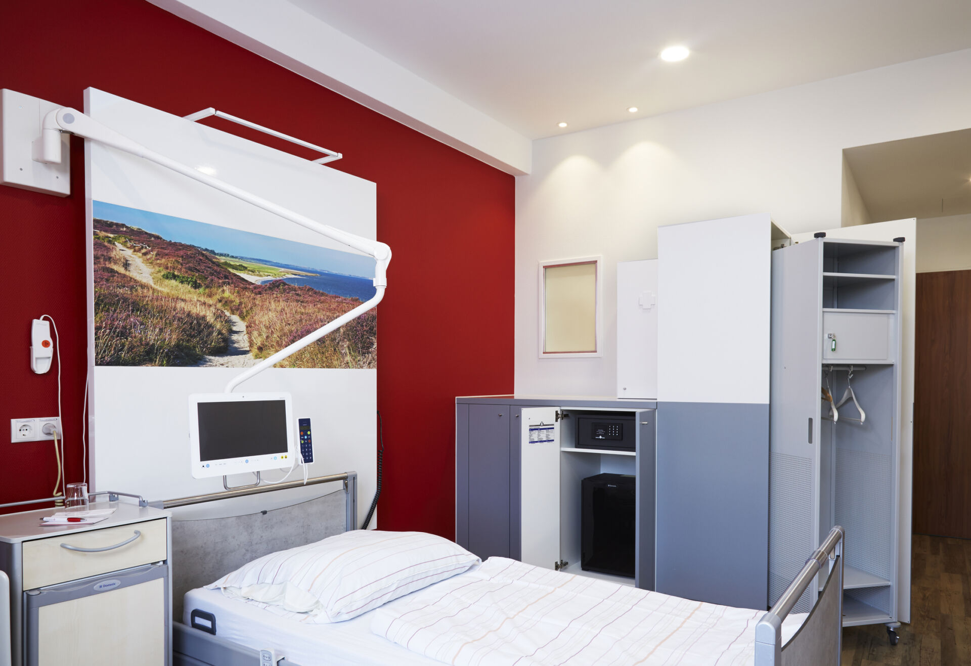 Modernes Krankenhauszimmer mit einem Bett, Schrank, Monitor und einem großen Bild an einer dunkelroten Wand.