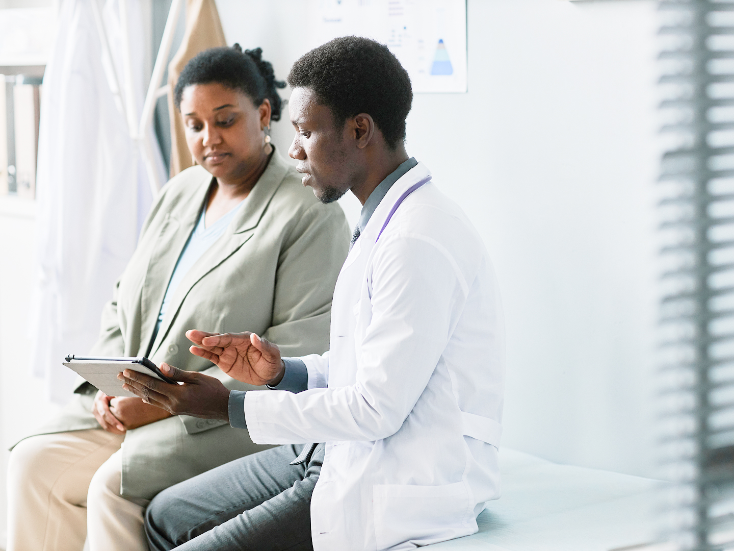 Ein Arzt im weißen Kittel erklärt einer Patientin etwas auf einem Tablet in einer hellen Arztpraxis.