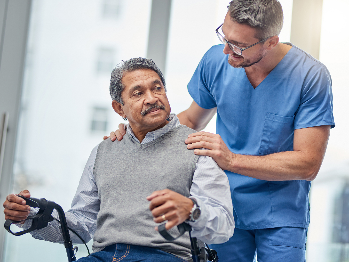Ein älterer Mann sitzt auf einem Rollator, während ein junger Arzt ihm freundlich von hinten auf die Schultern fasst.
