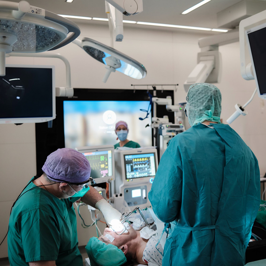 Chirurgisches Team in einem Operationssaal, das an einem Patienten arbeitet, umgeben von medizinischen Geräten.