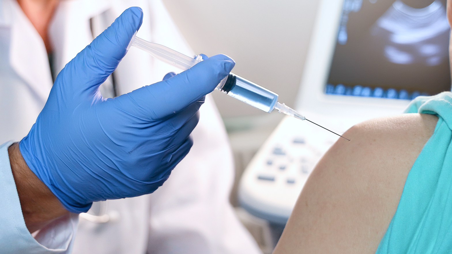 Arzt mit blauen Handschuhen hält eine Spritze an den Arm eines Patienten. Im Hintergrund ist ein Ultraschallgerät zu sehen.