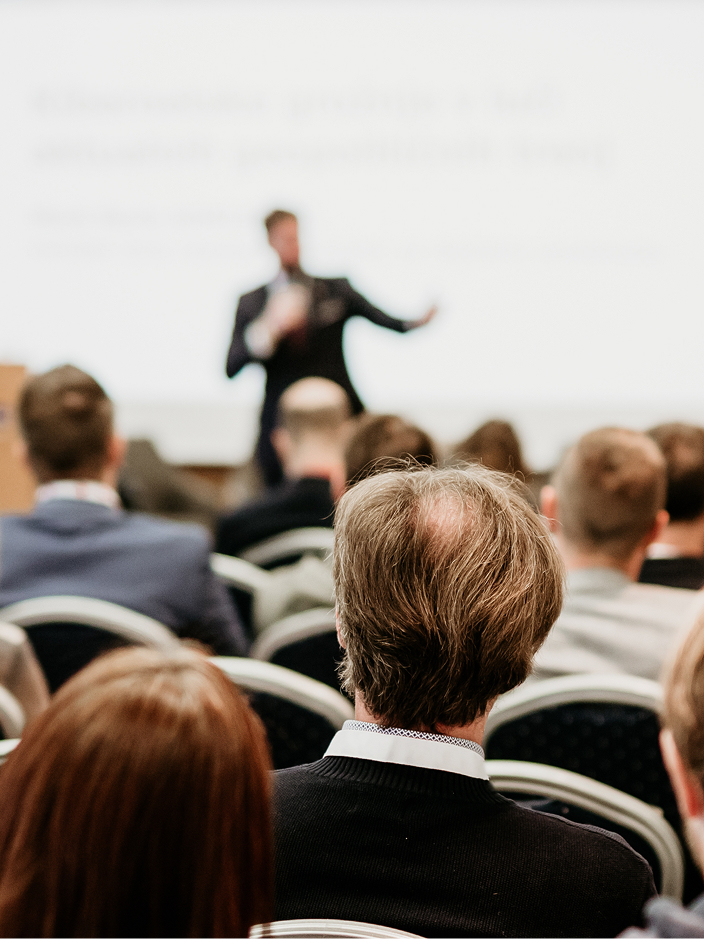 Ein Redner steht im Hintergrund vor einem Publikum in einem Konferenzraum, im Vordergrund sieht man die Rücken der Zuhörer.