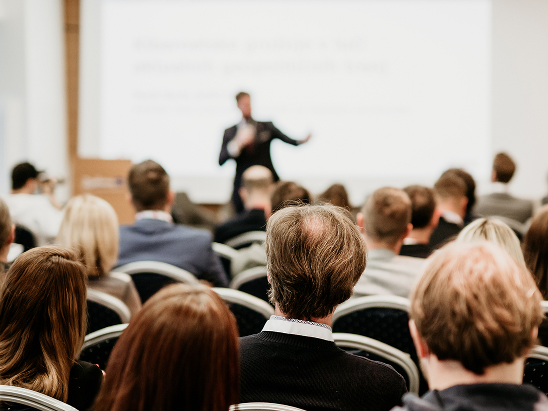 Ein Redner steht im Hintergrund vor einem Publikum in einem Konferenzraum, im Vordergrund sieht man die Rücken der Zuhörer.