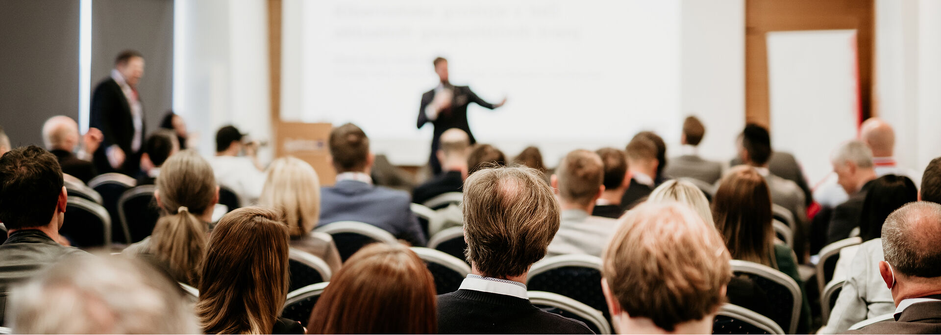 Ein Redner steht im Hintergrund vor einem Publikum in einem Konferenzraum, im Vordergrund sieht man die Rücken der Zuhörer.