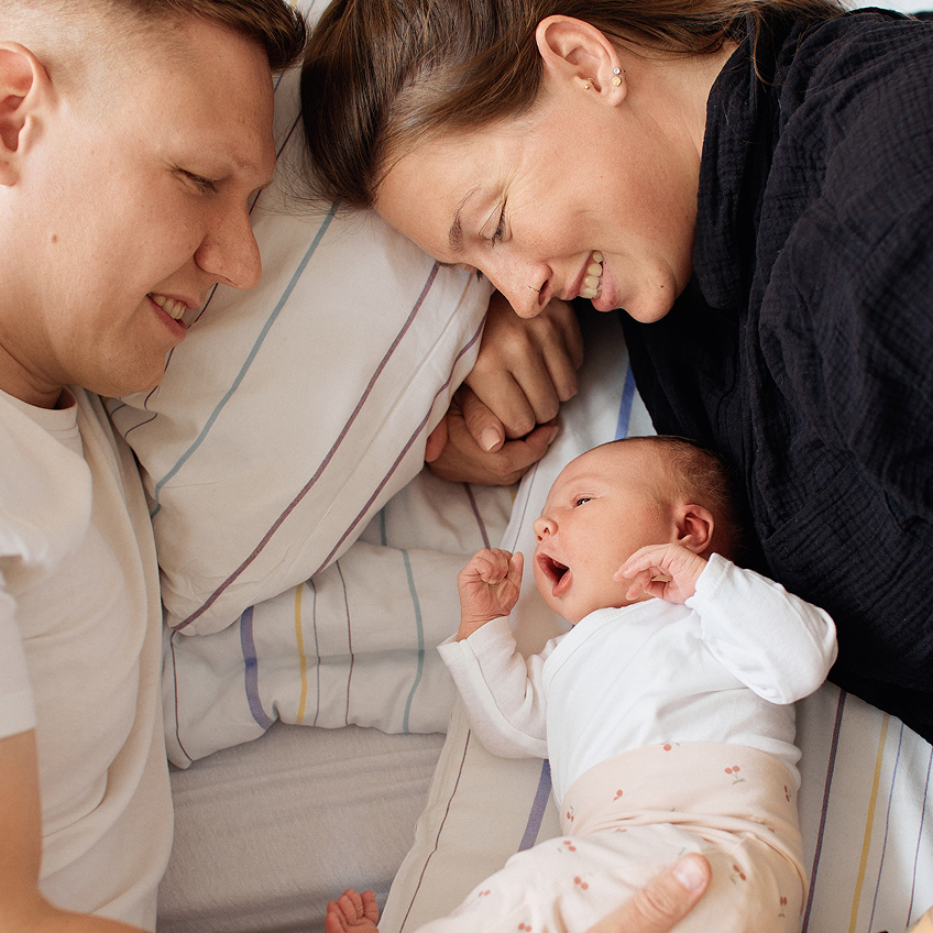 Ein Elternpaar liegt im Bett und schaut liebevoll auf ein Baby, dass zwischen ihnen liegt.
