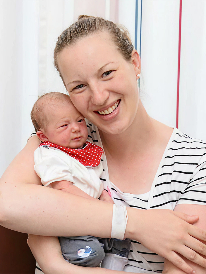 Lächelnde, blonde Mutter hält ihr Baby im Arm und schaut in die Kamera.