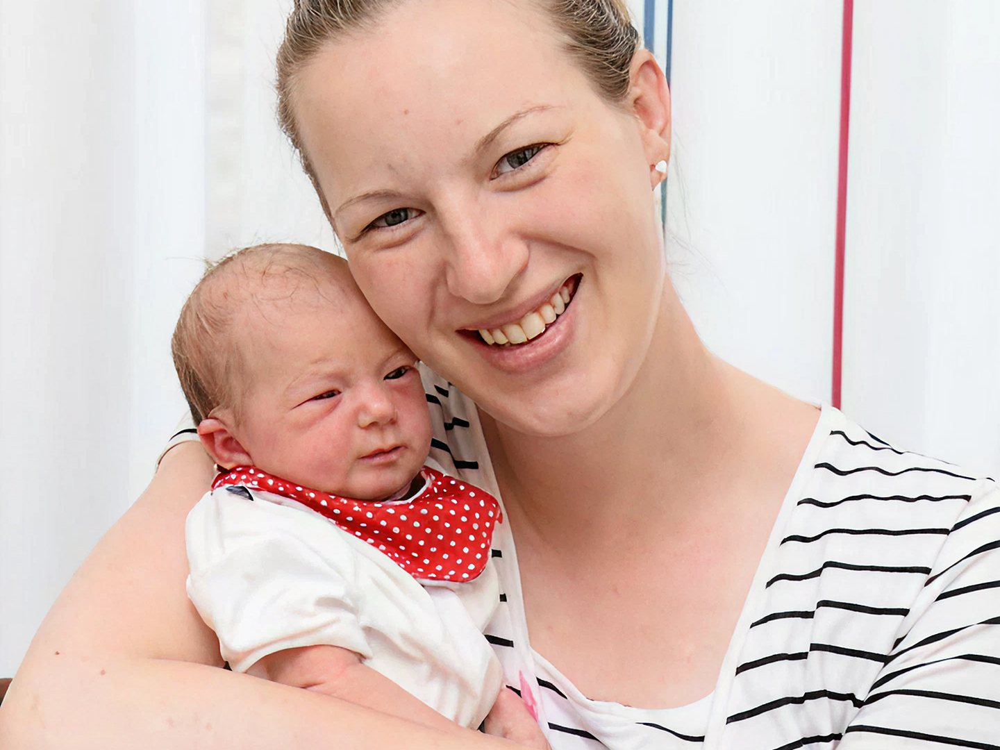 Lächelnde, blonde Mutter hält ihr Baby im Arm und schaut in die Kamera.