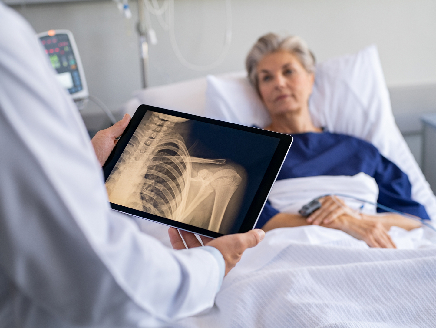 Ein Arzt im Vordergrund hält ein Tablet, das eine Röntgenaufnahme zeigt, im Hintergrund sieht man eine Patientin im Bett, die ihn ansieht.